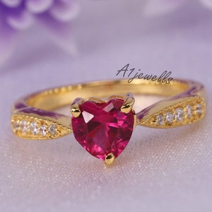 May include: A gold ring featuring a central heart-shaped red gemstone and small clear stones set along the band. The text A1jewells is visible above the ring. The band has a textured finish on the sides, leading to the prominent red heart center.