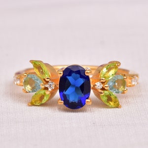 May include: A gold ring featuring a central oval-cut deep blue gemstone. The setting is flanked by marquise-cut light green and pale blue stones arranged in a leaf-like pattern, accented with small clear round stones on a polished gold band.