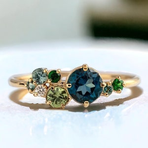 Può includere: Un anello d'oro con un gruppo di pietre preziose blu, verdi e bianche. La pietra più grande è una pietra rotonda blu al centro.