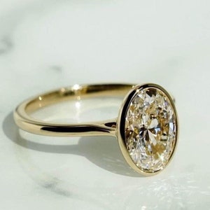 Puede incluir: Un anillo de compromiso de oro con engaste de bisel que presenta un gran diamante de forma ovalada.