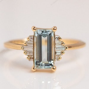 May include: A gold ring with a light blue emerald-cut aquamarine gemstone surrounded by smaller diamonds in a baguette setting.