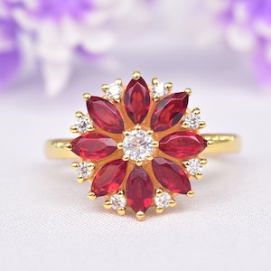 May include: A gold ring featuring a flower design with red marquise-cut gemstones and small white gemstones. The center of the flower has a single round white gemstone. The ring is set against a soft white background.