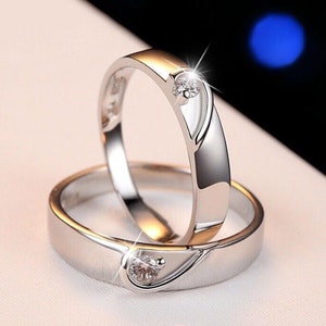 May include: Two silver rings with a heart-shaped design and a small, sparkling gemstone. The rings are displayed on a white surface, with a blurred background. The rings have a polished finish.