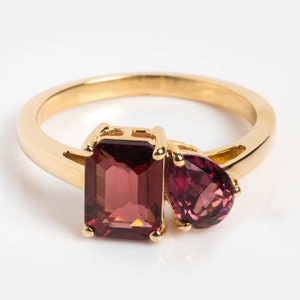 May include: A gold ring featuring two red gemstones. The larger stone is a rectangular emerald cut, and the smaller stone is a pear shape.