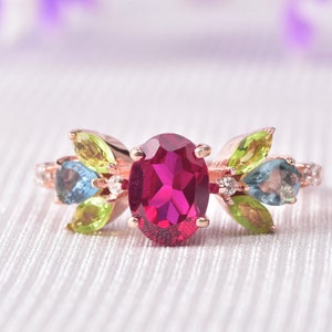May include: A rose gold ring with a large oval ruby gemstone surrounded by smaller green and blue gemstones.