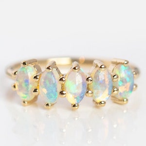 May include: A gold band ring with five oval-shaped opals. The opals are iridescent with shades of white, blue, and green.
