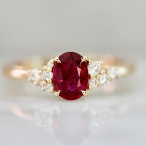May include: A gold ring featuring a large oval ruby center stone. The ruby is a deep red color and is surrounded by smaller, clear accent stones. The ring has a delicate, elegant design.