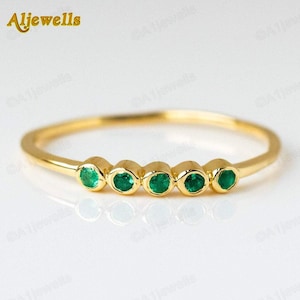 May include: A delicate gold ring featuring five round, vibrant green emeralds set in a row. The ring has a thin band and a polished, reflective surface. The brand name "Aljewells" is in the top left corner.
