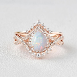 May include: Rose gold ring set with a pear-shaped opal gemstone and a halo of small, clear gemstones. The band has a twisted design with more small gemstones. The set includes a matching band with a crown-like design.