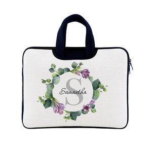 May include: A personalised laptop sleeve with a floral design. The white sleeve has a navy blue handle and trim, with a wreath of green leaves and purple flowers around a grey letter "S" and the name "Samantha".