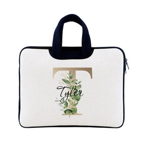 May include: White laptop sleeve with a navy blue handle and trim. The design features a gold letter "T" with a floral arrangement and the name "Tyler" in script. The sleeve has a zipper closure.