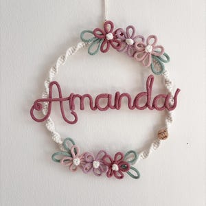 Può includere: Una ghirlanda in macramè fatta a mano con il nome "Amanda" in filato rosa. La ghirlanda è decorata con fiori in filato rosa, verde e bianco. Una piccola etichetta di legno è attaccata alla ghirlanda.