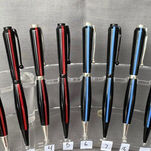 May include: Twelve black and silver pens with red and blue stripes. The pens are arranged in a row on a clear acrylic display stand. The numbers 1 through 12 are printed on the stand.