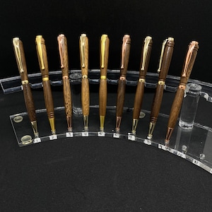 May include: Ten wooden pens with gold and copper accents displayed on a clear acrylic stand. The pens are arranged in a semi-circle with the numbers 1 through 11 visible on the stand.
