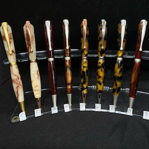 May include: Ten pens displayed on a clear acrylic stand. The pens are made of different materials, including wood, metal, and plastic. The pens are all different colors, including brown, black, white, and red. The pens are all different lengths and have different designs.