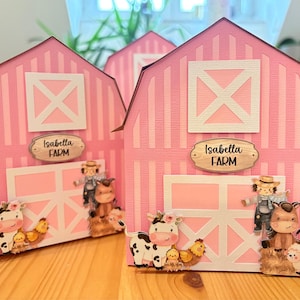 Può includere: Scatole regalo rosa a forma di fienile con dettagli bianchi e la scritta "Isabella FARM". Ogni scatola presenta una mucca, dei polli, un contadino, un cavallo e una pecora. Le scatole sono su una superficie di legno.