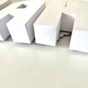 May include: Close-up of white paper letters spelling a word. The letters are three-dimensional and have a clean, modern design. The letters are arranged on a white surface, creating a minimalist aesthetic. The letters are likely used for decoration.
