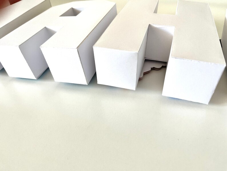 May include: Close-up of white paper letters spelling a word. The letters are three-dimensional and have a clean, modern design. The letters are arranged on a white surface, creating a minimalist aesthetic. The letters are likely used for decoration.