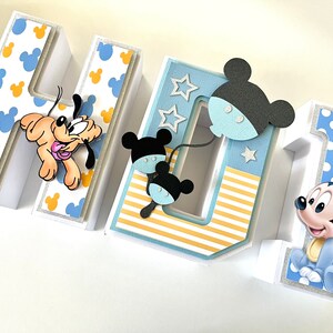 May include: A decorative, three-dimensional sign spelling out "NAME 1" with a Disney theme. The letters are white with blue and yellow Mickey Mouse patterns. Pluto and Mickey Mouse figures are attached to the letters. The number 1 has a blue polka dot pattern.