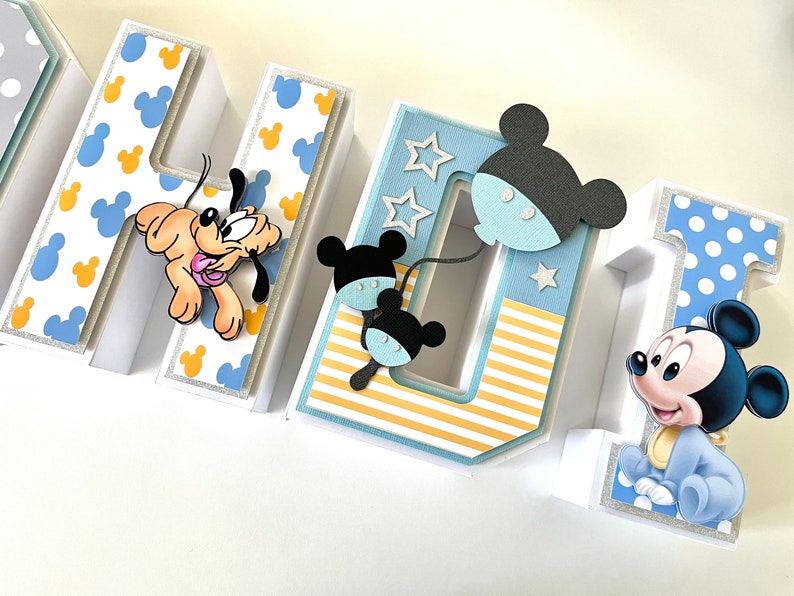 May include: A decorative, three-dimensional sign spelling out "NAME 1" with a Disney theme. The letters are white with blue and yellow Mickey Mouse patterns. Pluto and Mickey Mouse figures are attached to the letters. The number 1 has a blue polka dot pattern.