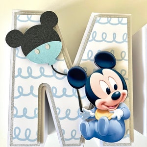 May include: A decorative letter "M" with a baby Mickey Mouse theme. The letter is white with a light blue squiggly pattern and silver glitter trim. A black and blue Mickey Mouse balloon and a baby Mickey Mouse are attached to the letter.