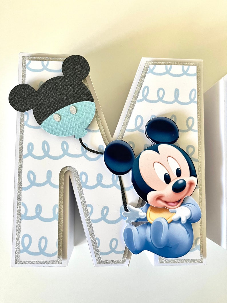 May include: A decorative letter "M" with a baby Mickey Mouse theme. The letter is white with a light blue squiggly pattern and silver glitter trim. A black and blue Mickey Mouse balloon and a baby Mickey Mouse are attached to the letter.