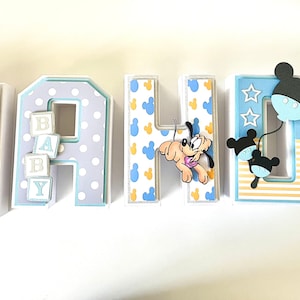 May include: A decorative set of letters spelling "MAHJI" with a baby theme. Each letter is decorated with Mickey Mouse and Pluto characters, polka dots, and baby blocks. The letters are in shades of blue, white, and yellow.