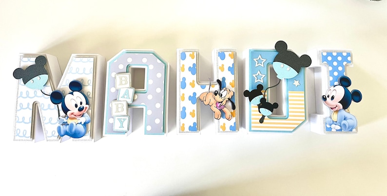 May include: A decorative set of letters spelling "MAHJI" with a baby theme. Each letter is decorated with Mickey Mouse and Pluto characters, polka dots, and baby blocks. The letters are in shades of blue, white, and yellow.