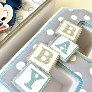 May include: A handmade baby-themed decorative box set. The boxes are white and light blue with polka dots and Mickey Mouse accents. The word "BABY" is spelled out with individual letter blocks. The boxes are decorated with silver glitter.