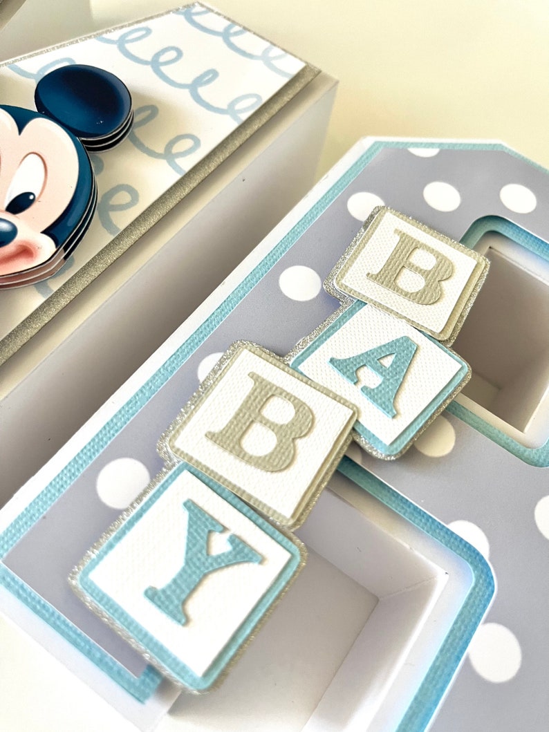May include: A handmade baby-themed decorative box set. The boxes are white and light blue with polka dots and Mickey Mouse accents. The word "BABY" is spelled out with individual letter blocks. The boxes are decorated with silver glitter.