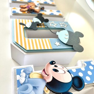 May include: A decorative papercraft featuring the name "Babe" in layered, 3D letters. Each letter is adorned with cartoon character cutouts, including Mickey Mouse and a teddy bear, and accented with blue, yellow, and white patterns. The letters are set against a white background.