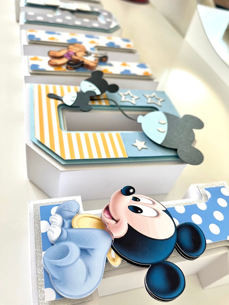 May include: A decorative papercraft featuring the name "Babe" in layered, 3D letters. Each letter is adorned with cartoon character cutouts, including Mickey Mouse and a teddy bear, and accented with blue, yellow, and white patterns. The letters are set against a white background.