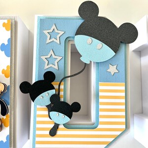 May include: A decorative letter "O" with a Mickey Mouse theme. The letter is light blue with yellow and white stripes. Mickey Mouse cutouts in black and light blue are attached to the letter, along with silver star accents.