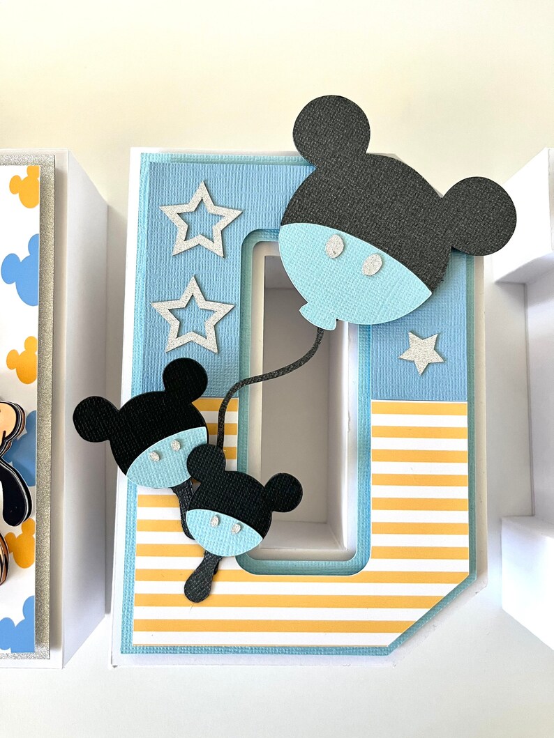 May include: A decorative letter "O" with a Mickey Mouse theme. The letter is light blue with yellow and white stripes. Mickey Mouse cutouts in black and light blue are attached to the letter, along with silver star accents.