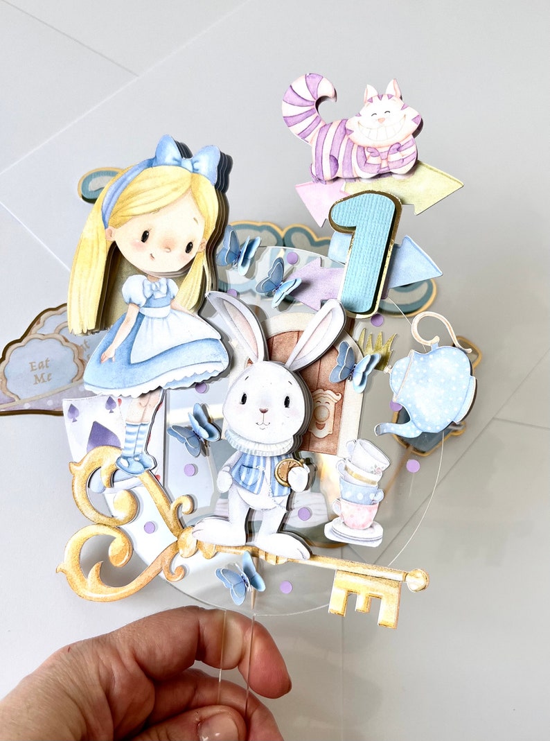 Shown here is an Alice in Wonderland-inspired cake decoration, mounted on a clear acrylic circle with the characters drawn in watercolor. Made with high quality materials