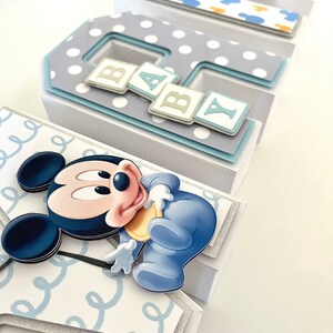 May include: A decorative baby-themed display featuring the letters B, A, B, and Y. Each letter is a layered box with a different design, including polka dots, Mickey Mouse, and a baby-themed Mickey Mouse. The letters are in shades of blue, white, and gray.