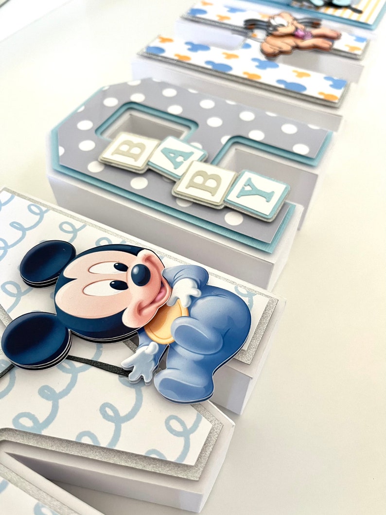 May include: A decorative baby-themed display featuring the letters B, A, B, and Y. Each letter is a layered box with a different design, including polka dots, Mickey Mouse, and a baby-themed Mickey Mouse. The letters are in shades of blue, white, and gray.