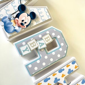 May include: Decorative paper letters spelling "BABY" with a Mickey Mouse theme. The letters are white, gray, and blue, with polka dots, Mickey Mouse silhouettes, and glitter accents. A Mickey Mouse figure and Pluto are attached to the letters.