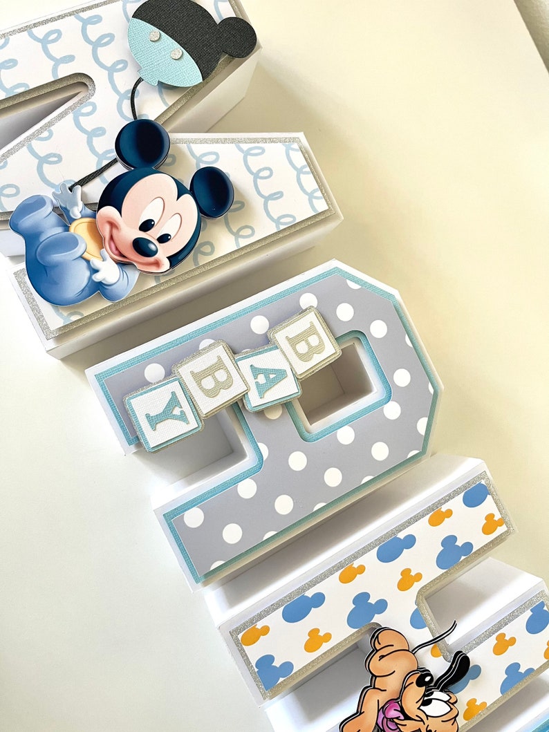 May include: Decorative paper letters spelling "BABY" with a Mickey Mouse theme. The letters are white, gray, and blue, with polka dots, Mickey Mouse silhouettes, and glitter accents. A Mickey Mouse figure and Pluto are attached to the letters.