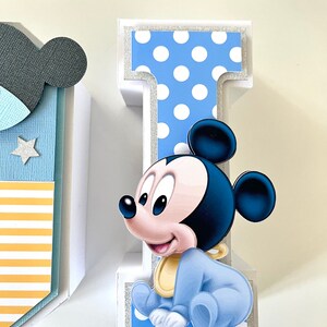 May include: A decorative letter "I" with a baby Mickey Mouse design. The letter is white with a blue polka dot pattern and silver glitter accents. Mickey Mouse is wearing a blue outfit and a yellow bib.