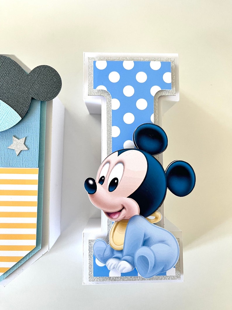 May include: A decorative letter "I" with a baby Mickey Mouse design. The letter is white with a blue polka dot pattern and silver glitter accents. Mickey Mouse is wearing a blue outfit and a yellow bib.