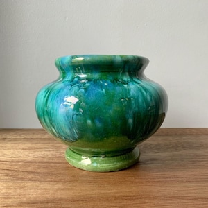 May include: A small, round ceramic vase with a wide opening and a pedestal base. The vase is glazed in shades of green and blue, creating a marbled effect. The vase is sitting on a wooden surface.
