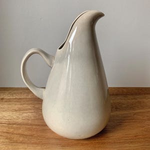 May include: A cream-coloured ceramic jug with a curved handle and a spout. The jug has a smooth, glossy finish and a teardrop shape. It is on a wooden surface.