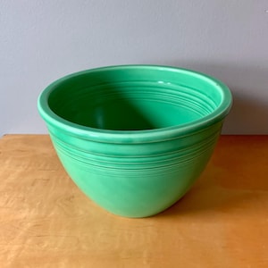 May include: A vibrant green ceramic bowl with a rounded shape and a wide rim. The bowl has horizontal ridges around the exterior, adding texture to the design. The bowl is sitting on a wooden surface.