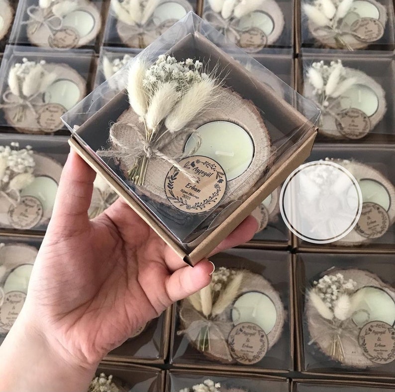 K&ouml;nnte beinhalten: Eine quadratische Geschenkbox aus Karton mit einer nat&uuml;rlichen Holzscheibe, einem runden Teelicht, einem kleinen Strau&szlig; getrockneter wei&szlig;er Blumen und einem runden Papieranh&auml;nger, bedruckt mit den Namen Ayg&uuml;l und Erhan sowie dem Jahr 2021.