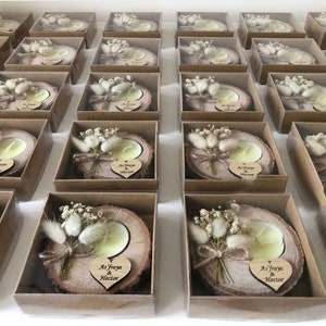 K&ouml;nnte beinhalten: Eine Sammlung von Teelichtern auf nat&uuml;rlichen Holzscheiben, jeweils verpackt in einer quadratischen braunen Pappschachtel. Die Kerzen sind mit getrockneten wei&szlig;en Blumen und einem kleinen Holzherz mit der Gravur As'fraya & Hector verziert.