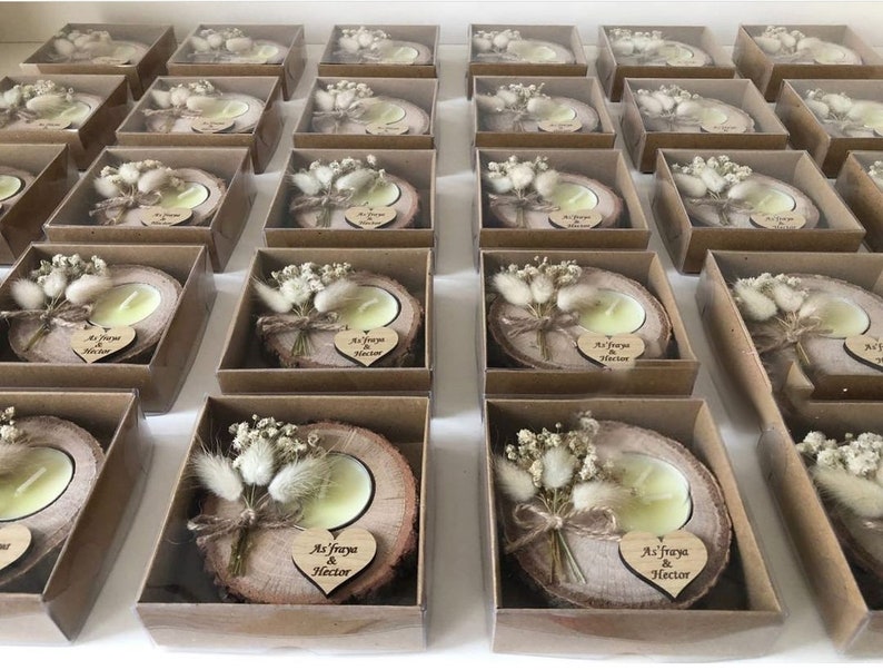 K&ouml;nnte beinhalten: Eine Sammlung von Teelichtern auf nat&uuml;rlichen Holzscheiben, jeweils verpackt in einer quadratischen braunen Pappschachtel. Die Kerzen sind mit getrockneten wei&szlig;en Blumen und einem kleinen Holzherz mit der Gravur As'fraya & Hector verziert.