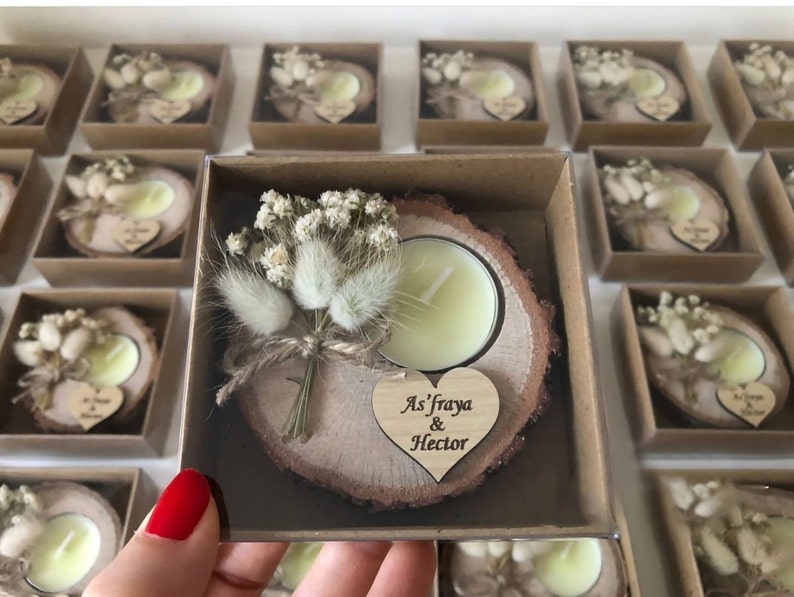 May include: A round wooden slice candle holder with a light yellow tea light candle, decorated with a small bouquet of dried white flowers and grasses, and a heart-shaped wooden tag engraved with the text As'fraya & Hector in a box.
