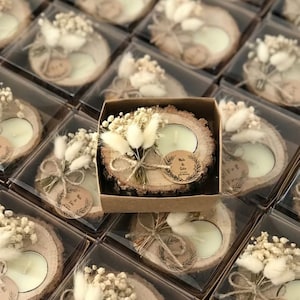 May include: A round wood slice candle holder with bark edges, featuring a white tea light candle, a small bouquet of dried white flowers and bunny tails, and a circular paper tag with the printed text Melis & Selin, all inside a square box.