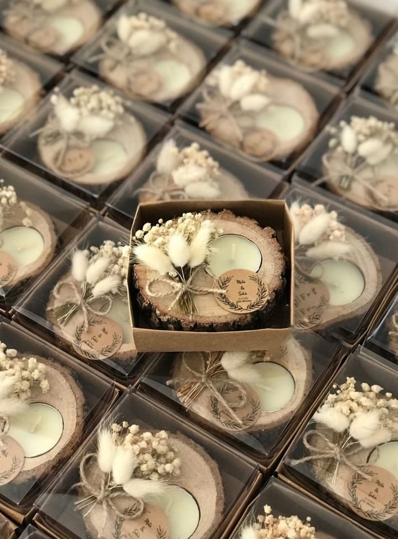 May include: A round wood slice candle holder with bark edges, featuring a white tea light candle, a small bouquet of dried white flowers and bunny tails, and a circular paper tag with the printed text Melis & Selin, all inside a square box.